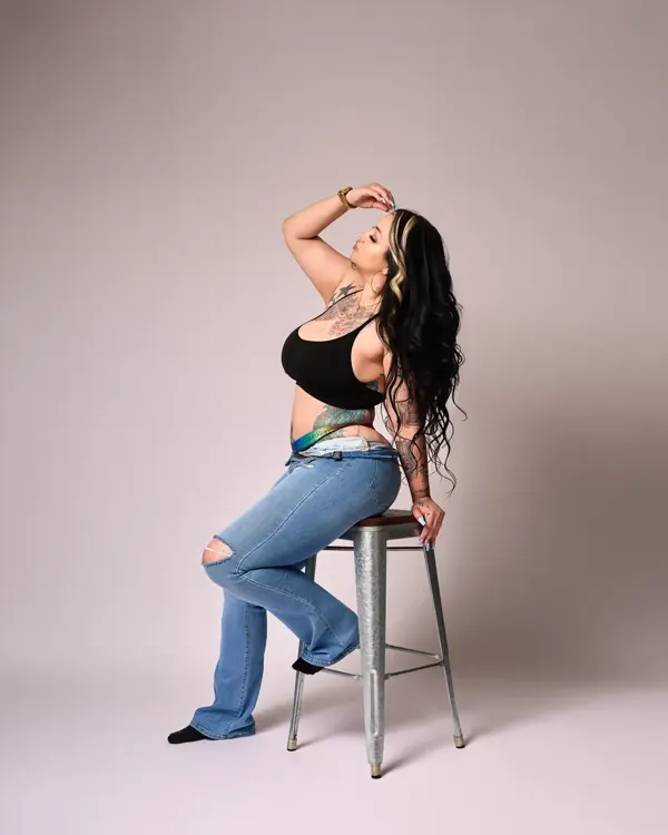 Khloe Lynn in a black top and ripped jeans perched on a studio stool