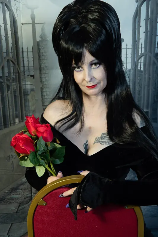 Selina Dark in black velvet holding red roses, seated in a red velvet chair