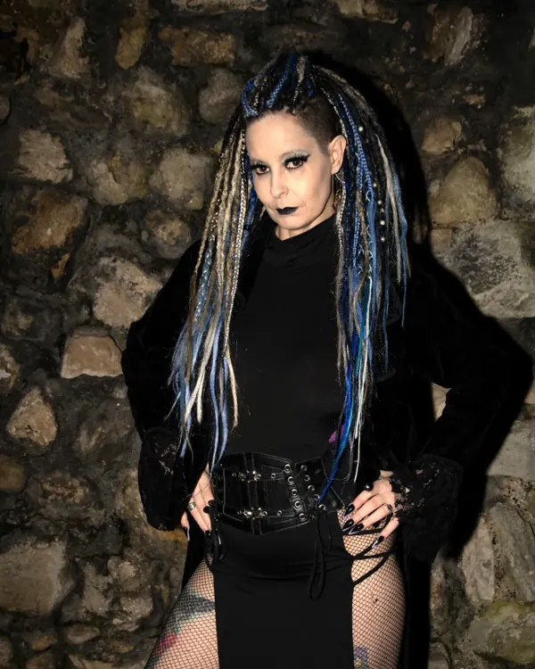 Selina Dark with blue-streaked braids in a black outfit against a stone wall