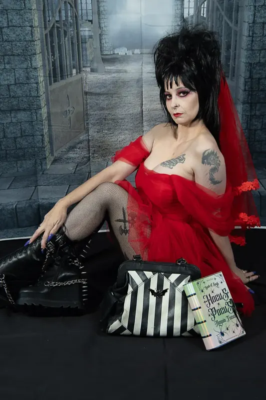 Selina Dark in a red dress and veil sitting beside a striped bag on a dark set